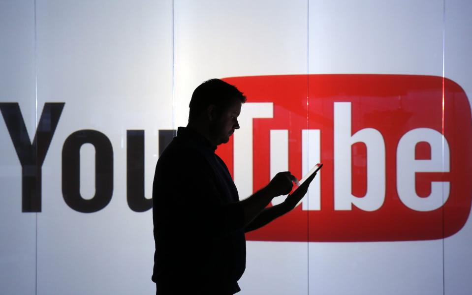 A man checks a mobile device while standing against an illuminated wall bearing YouTube's logo