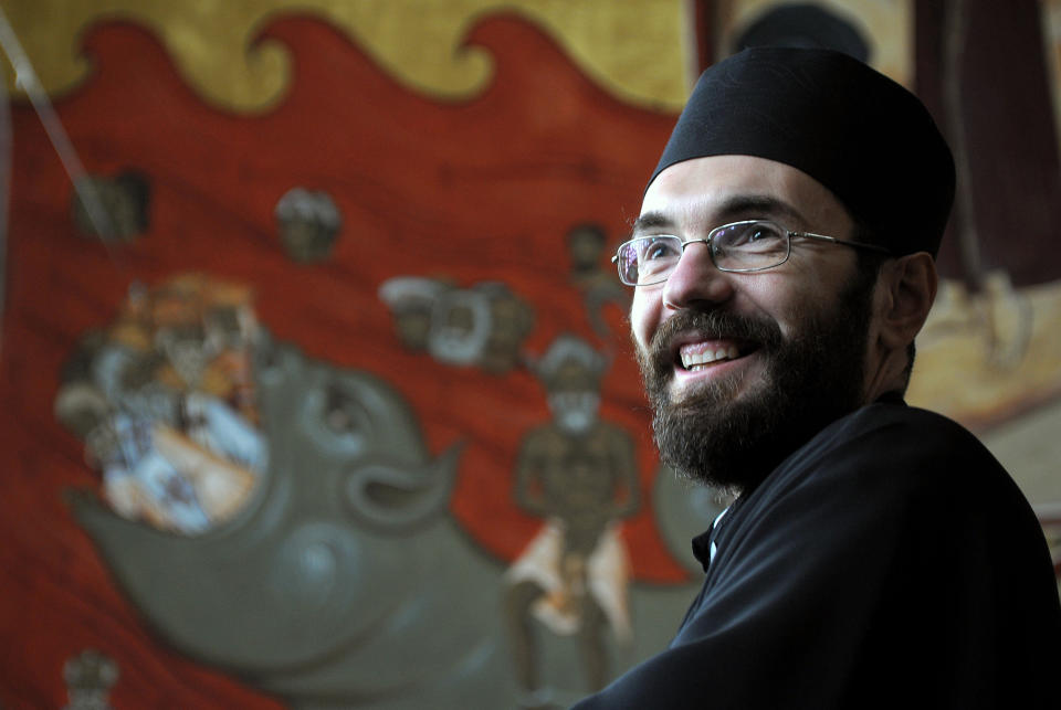In this photo taken Tuesday, Feb. 4, 2014 Orthodox priest Branko Vujacic smiles in front of the brightly-colored newly painted fresco in the Serbian Orthodox Church of Christ's Resurrection in Montenegro's capital Podgorica. The fresco allegedly shows late Yugoslav autocratic leader Josip Broz Tito drowning in red fiery waves of hell with Karl Marx and Friedrich Engels, authors of the 1848 Communist Manifesto. They are in the company of Adam and Eve, together with some of Montenegro's current politicians and people wearing Muslim turbans. What appear to be rival church priests are being swallowed through a huge jaw of an angry gray beast with pointed devil ears. The fresco has triggered much attention and public controversy in this tiny former communist country. (AP Photo/Risto Bozovic)