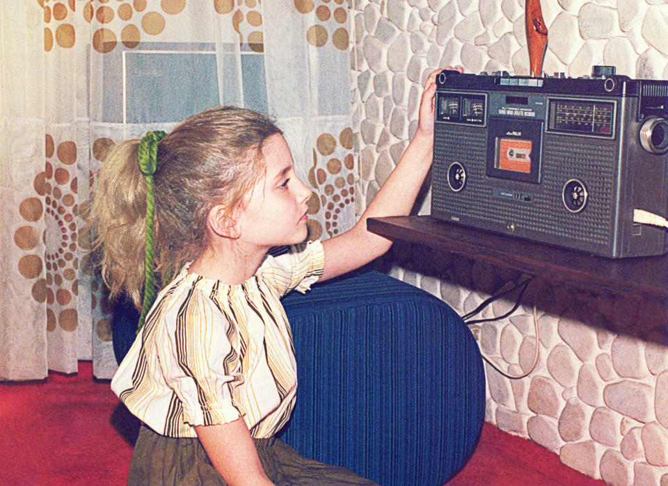 A young girl in vintage attire with her hair tied back listens to a cassette player in a retro-themed room with patterned curtains and textured walls