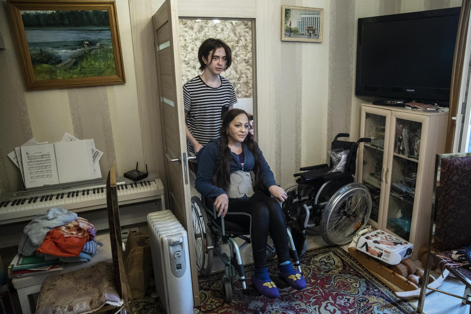 In this May 26, 2020, photo, Irina Karabulatova is pushed in a wheelchair by her son, Mikhail, in their apartment in Moscow, Russia. A smartphone app designed to track Moscow's quarantined coronavirus patients was rolled out by city officials in early April, but complaints about it have mushroomed. Karabulatova, a bed-ridden professor who hasn’t left her apartment in a year, was fined for not installing the mandatory app. After her story made national headlines, the fines were canceled and officials apologized. (AP Photo/Pavel Golovkin)