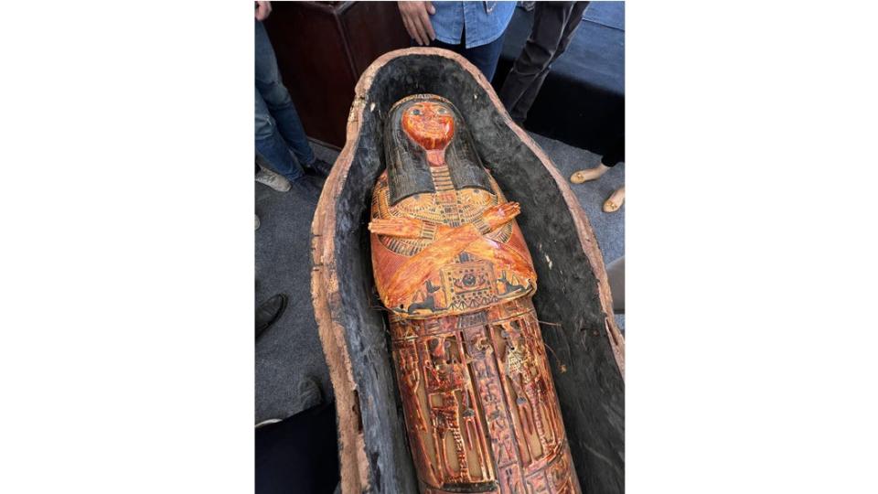 A top shot of an open sarcophagus shows an ornate ancient Egyptian cartonnage representing a woman with arms crossed across her chest.