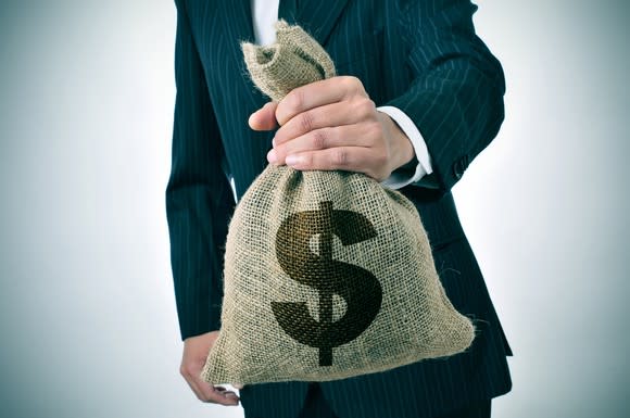 A man in a suit holds a burlap sack with a U.S. dollar sign emblazoned on it.