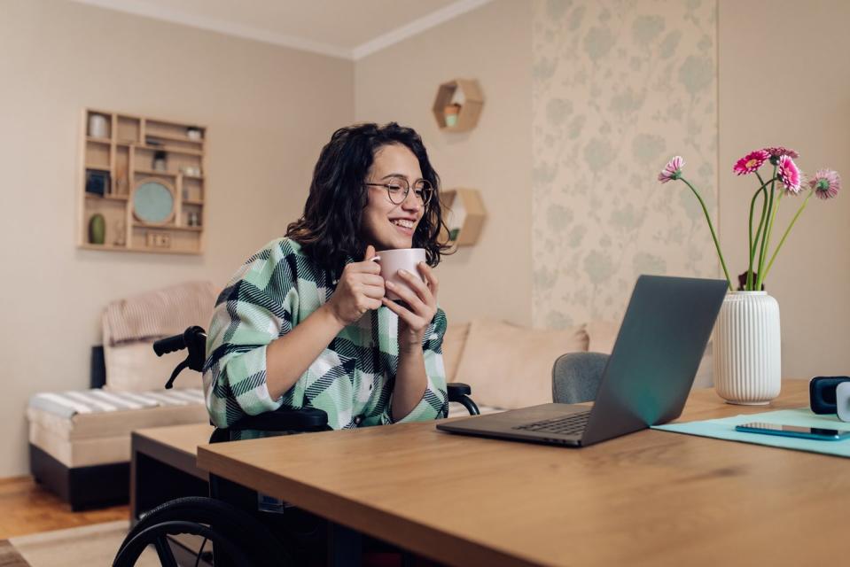 A person in a wheelchair who's holding a coffee mug and looking at an open laptop on a table in front of them.