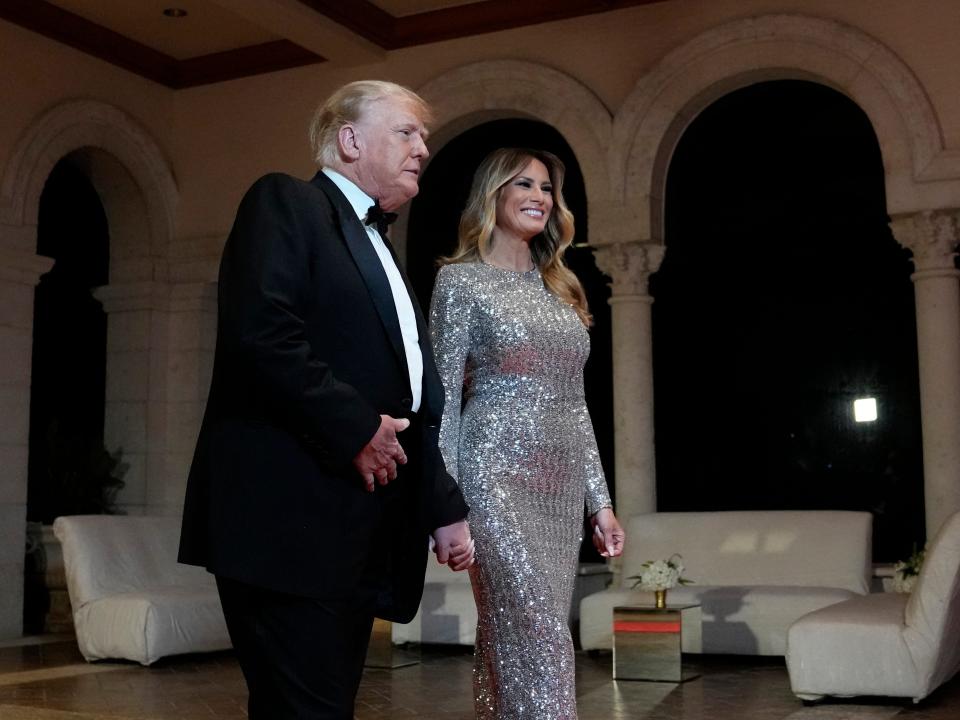 Former President Donald Trump in a black suit and bowtie holding hands with former first lady Melania Trump in a silver dress