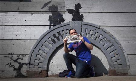 A man holds up a local newspaper as he poses for a photo in front of a new art piece by British graffiti artist Banksy in the Brooklyn borough of New York, October 17, 2013. REUTERS/Carlo Allegri