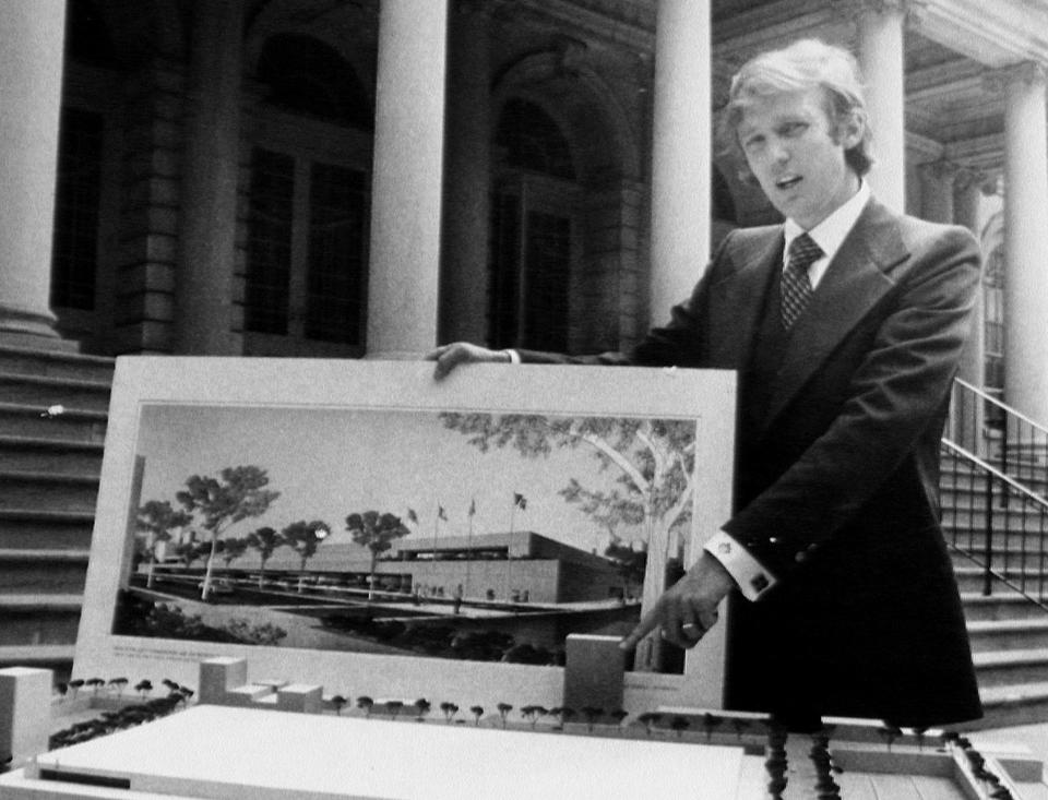 Donald Trump stands outdoors in front of a building, displaying a large architectural model and a rendering of a complex