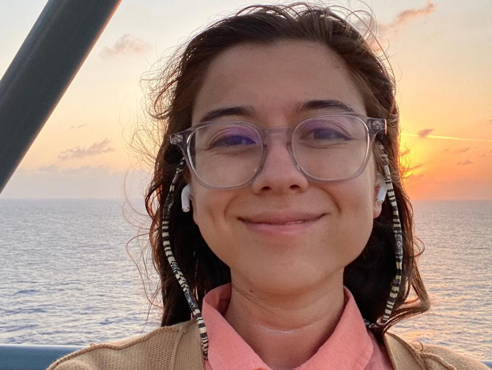 The author wears earbuds in front of the sun setting over the sea on the top deck of the cruiseship