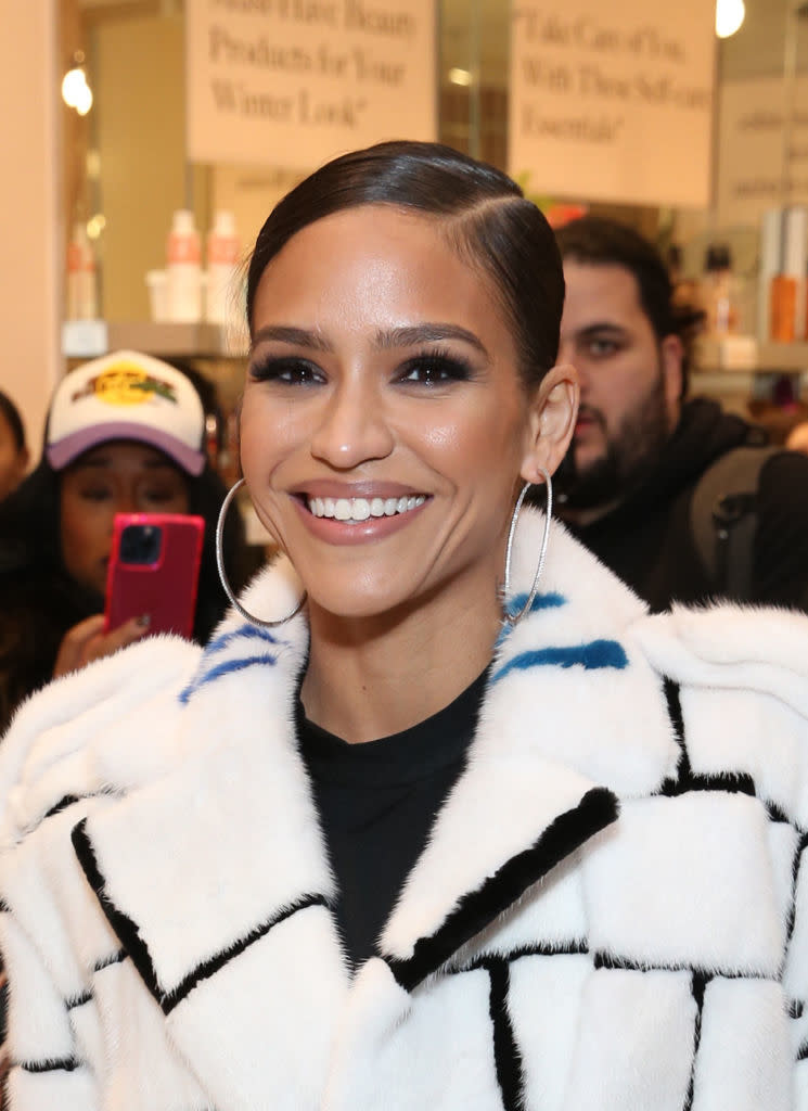 Cassie, with hoop earrings, a sleek hairstyle, and a stylish white coat with black patterns, smiles at an event. More people with a phone are in the background