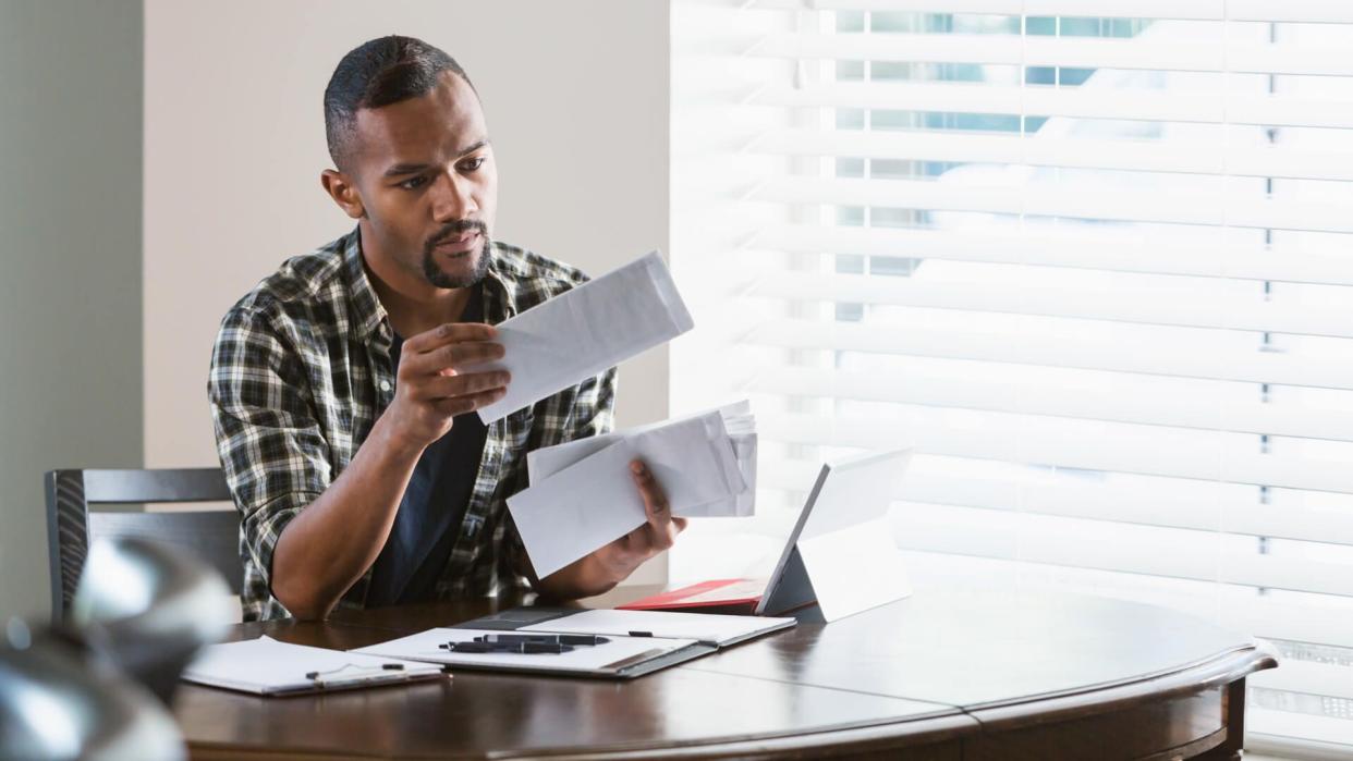 A young African-American man sitting at a desk by a window at home, paying bills.