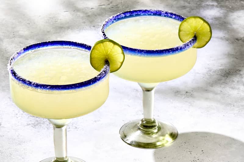 angled shot of two margaritas in large blue rimmed margarita glasses, garnished with lime wheels