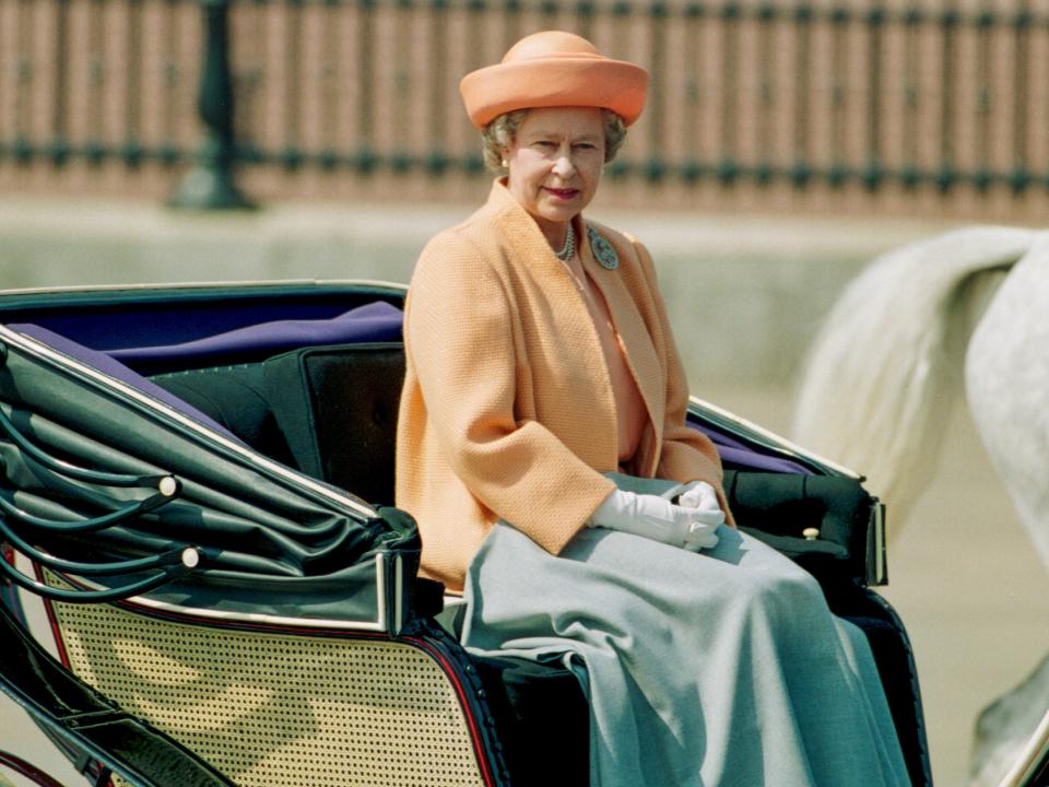 The Queen riding in an open carriage wearing a sherbert orange overcoat with a blanket over her legs. She has on round matching hat and white gloves.