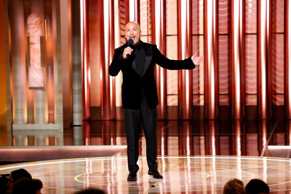 A smiling man in a dark suit onstage holds a mic to his mouth and gestures with one hand