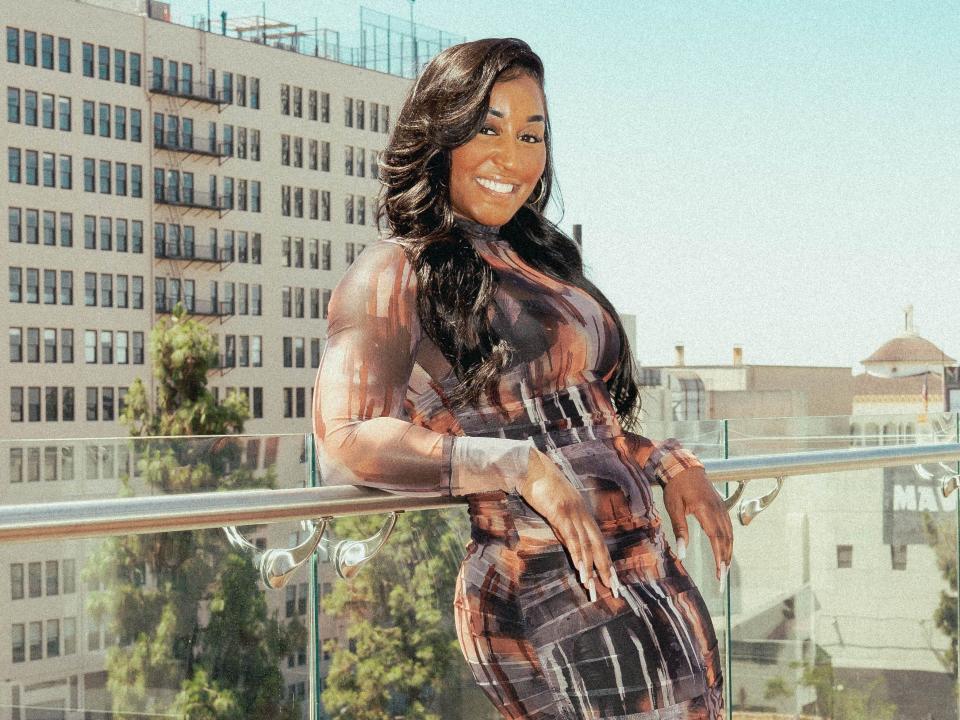 Choreographer Saleemah E. Knight wears a colorful mesh bodycon dress, stilettos, and hoop earrings while smiling into the camera. She's standing on a rooftop against a backdrop of an LA cityscape..
