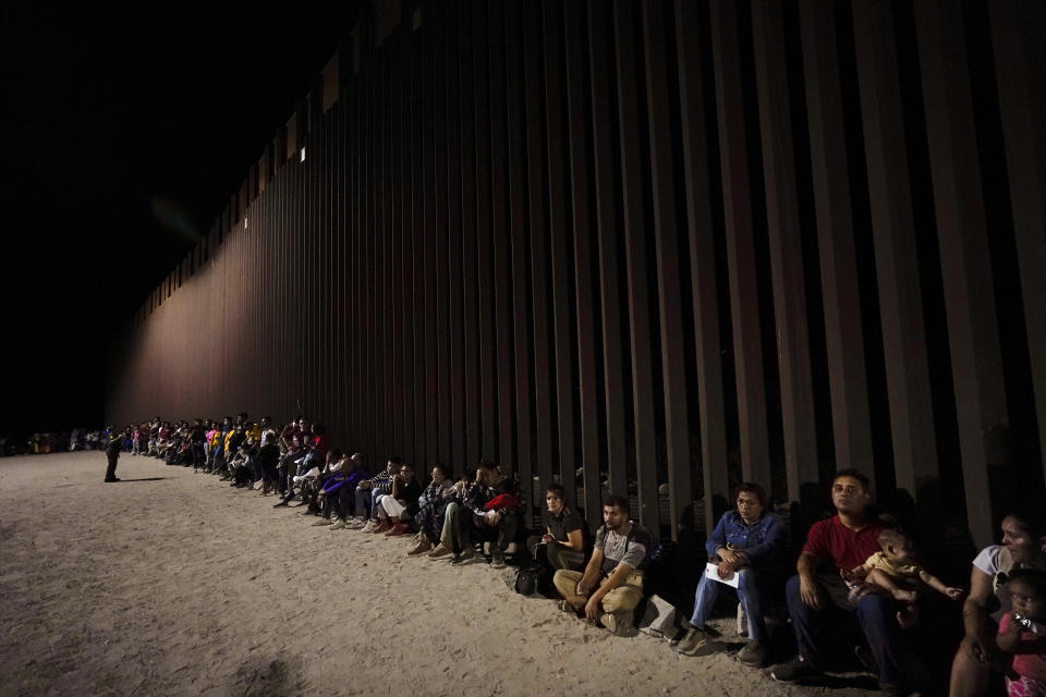 FILE - Migrants wait along a border wall Aug. 23, 2022, after crossing from Mexico near Yuma, Ariz. U.S. immigration offices have become so overwhelmed with processing migrants for court that some some asylum-seekers who crossed the border at Mexico may be waiting a decade before they even get a date to see a judge. The backlog stems from a change made two months after President Joe Biden took office, when Border Patrol agents began now-defunct practice of quickly releasing immigrants on parole. They were given instructions to report to a U.S. Immigration and Customs Enforcement office at their final destination to be processed for court — work previously done by the Border Patrol. (AP Photo/Gregory Bull, File)