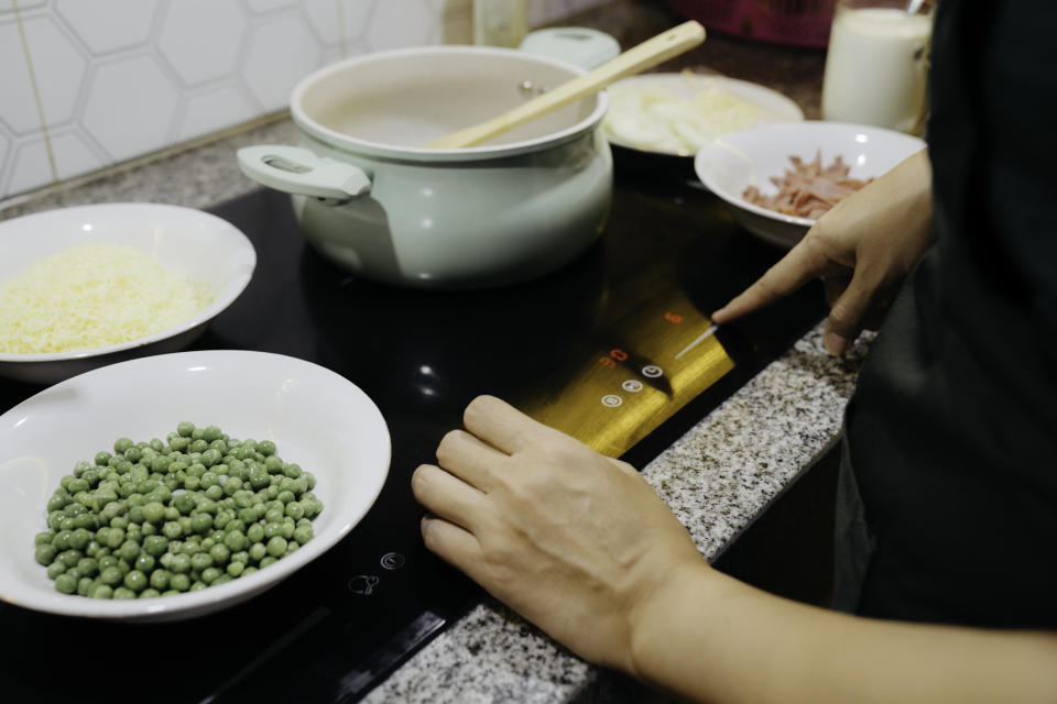 A person is using an induction cooktop while preparing ingredients, including peas, grated cheese, and chopped meat