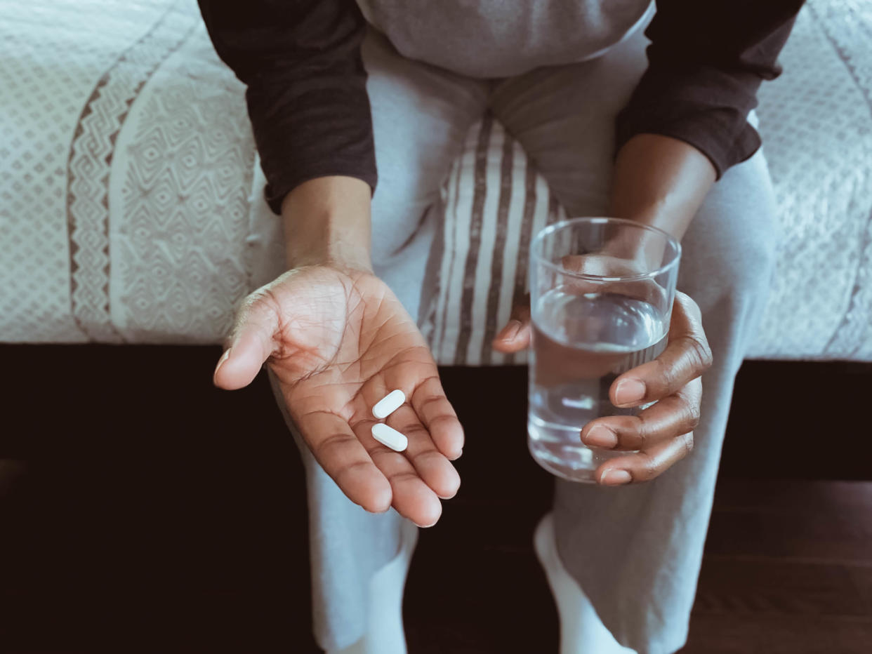 Frau sitzt auf Bettkante und hat zwei Tabletten und ein Glas Wasser in den Händen