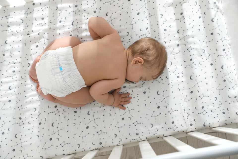 Baby sleeps on tummy in crib. The crib sheet has a celestial pattern of stars and moons. The baby is wearing a diaper