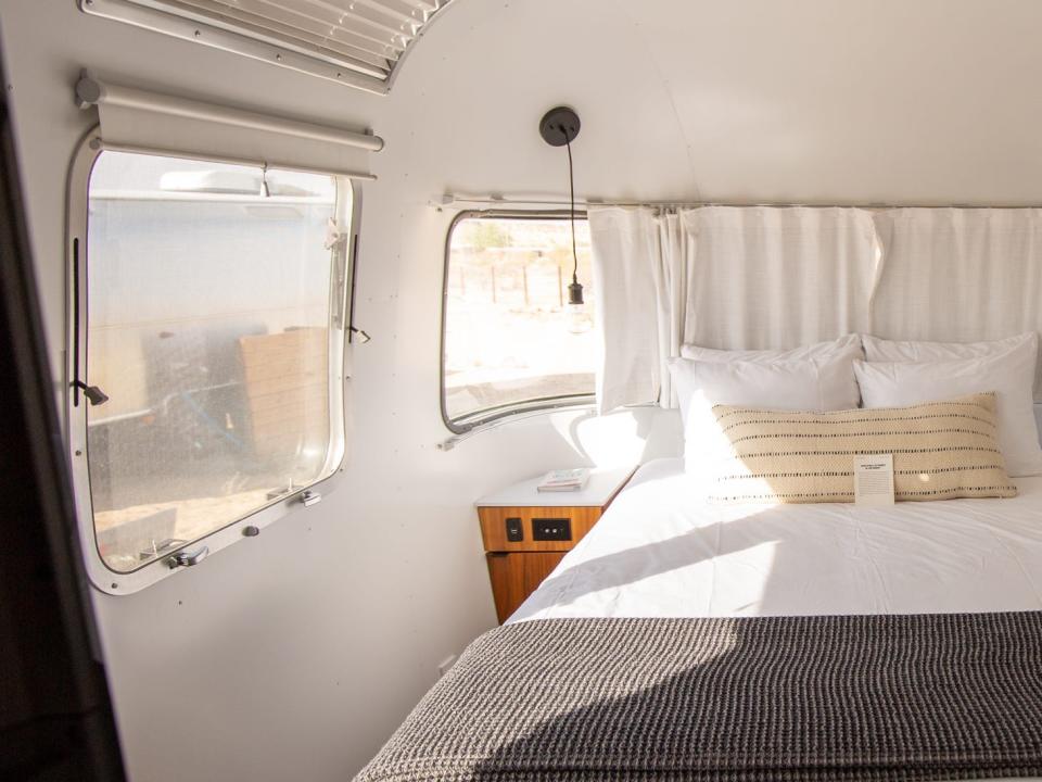 A bedroom inside Autocamp Joshua Tree's Airsteram trailer with white accents, a bed with pillows, curtains over windows.