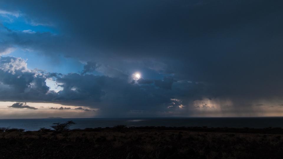 an eclipsed sun shines behind many storm clouds and rain is visible in the distance.