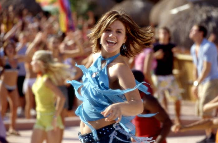Kelly in fringed top and belted denim shorts dancing with joy, others in background