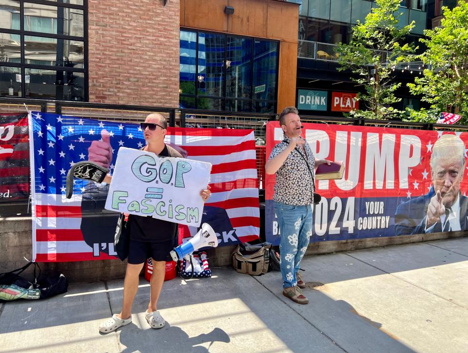 Denver resident Derek Torstenson, left, demonstrates against former President Donald Trump next to a man reading the Bible in front of Trump banner just outside the security perimeter at the Republican National Convention in Milwaukee on July 17, 2024.