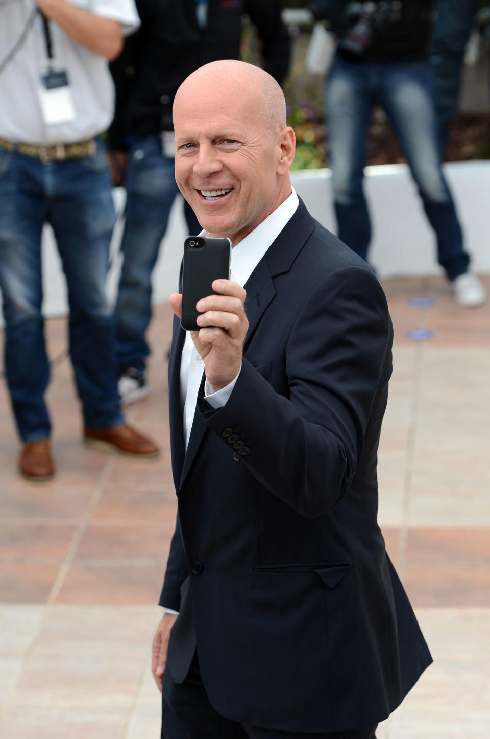 Bruce Willis on a red carpet, smiling and holding up a smartphone, wearing a suit with a shirt