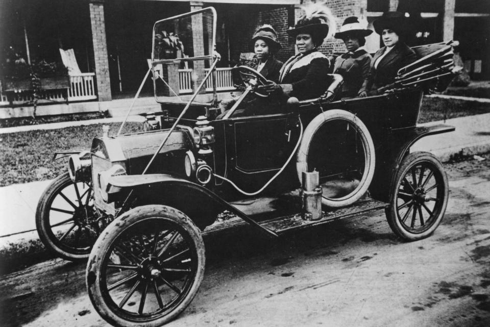 madam cj walker sits in the driver seat of an early car with the top down, a woman sits in the passenger seat, both women wear hats