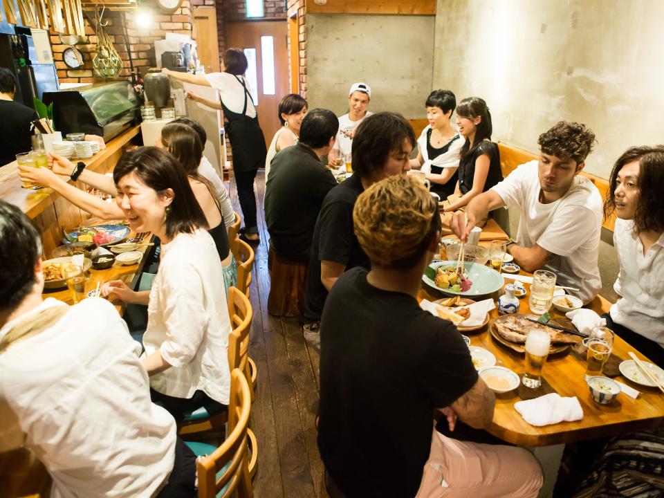 Several people sit at crowded tables in a warmly-lit Japanese eatery.