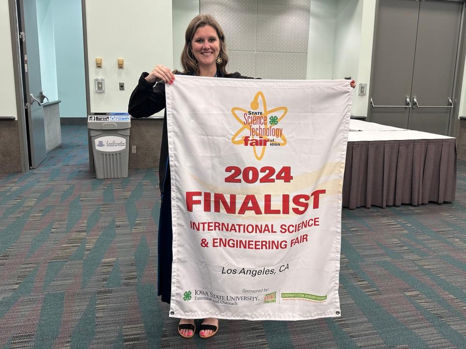 smiling woman in conference room hold up large white banner with red writing declaring 2024 FINALIST INTERNATIONAL SCIENCE & ENGINEERING FAIR