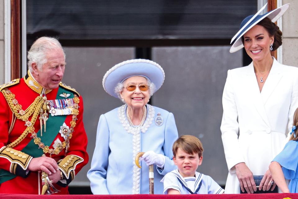 The Queen smiling in a pale blue coatdress with a white and silver petal design around her collar and down the center opening. She has on a matching blue and white wide-brimmed hat, pearls, an oval brooch, brown tinted sunglasses and a walking cane.