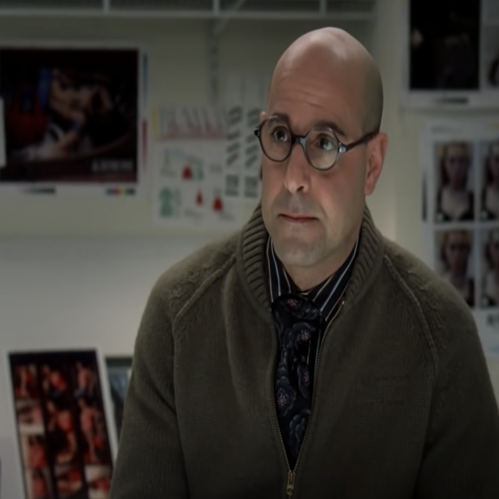 A bald man wearing round glasses and a sweater looks ahead