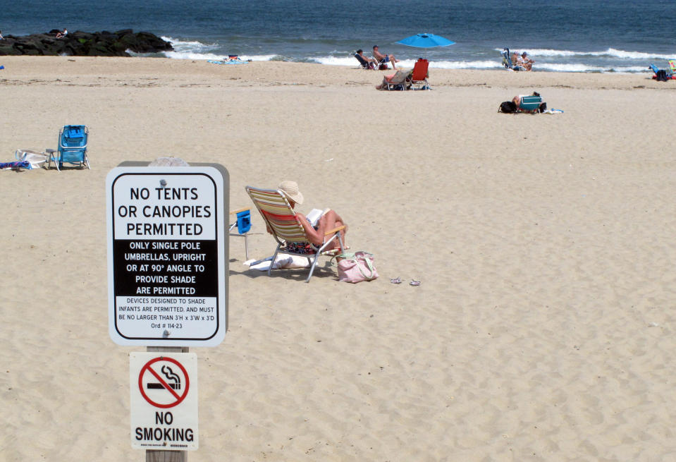In this Monday, May 20, 2019 photo, beachgoers sit on the sand beyond a sign indicating that smoking is prohibited on the beach in Spring Lake, N.J. A statewide smoking ban is in effect at New Jersey's beaches for the start of the 2019 summer season, although towns can set aside up to 15% as smoking sections. (AP Photo/Wayne Parry)