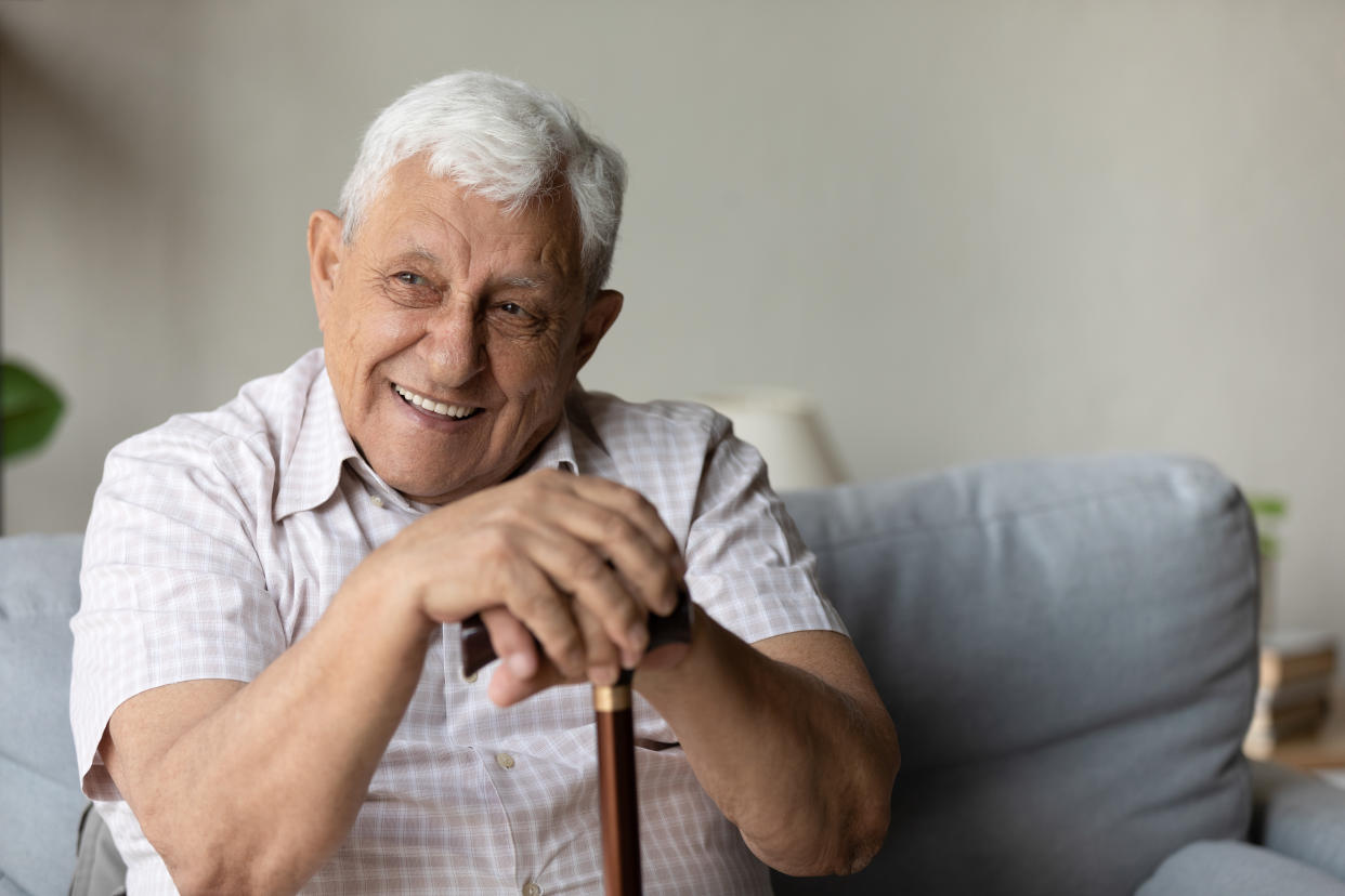 Happy dreamy senior elderly grandfather holding hands on walking wooden cane, resting on sofa. Joyful candid Old retired man looking in distance, enjoying carefree peaceful moment alone at home.