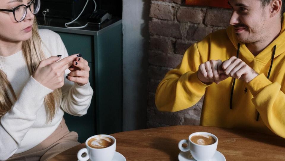 woman in glasses and white sweater sitting at a table with a man in an orange sweater signing ASL over coffee