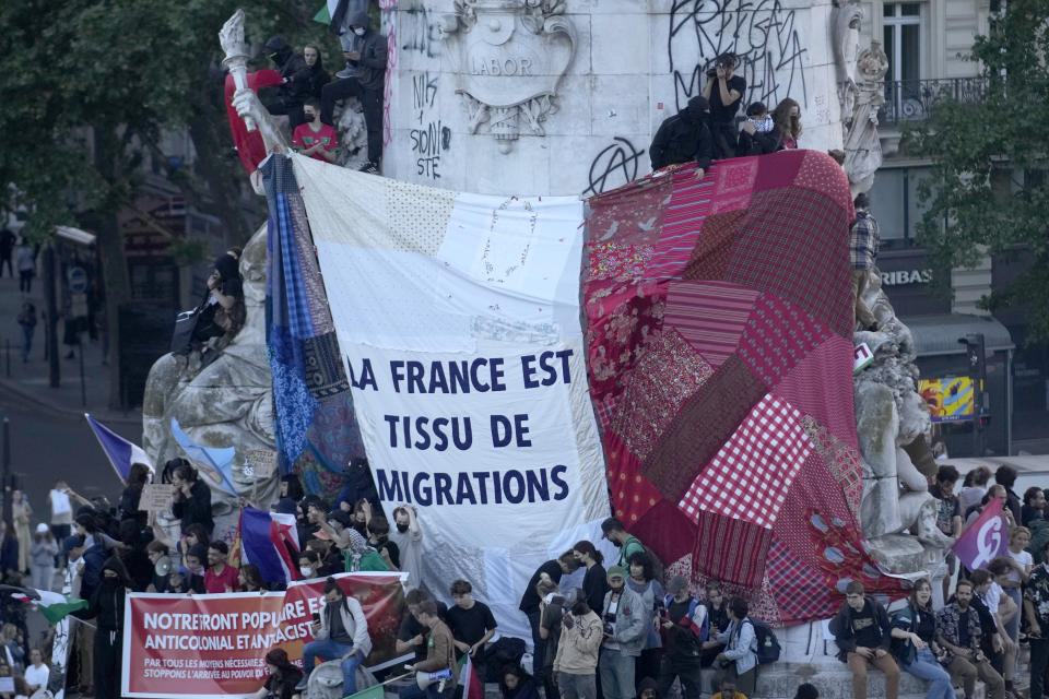 FILE - People stand near a French flag reading "France is a patchwork of migration" in Republique Plaza as they react to the projection of results during the second round of the legislative elections, in Paris, on July 7, 2024. (AP Photo/Christophe Ena, File)