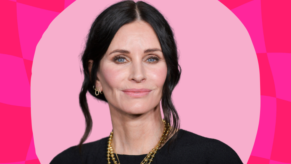 Courteney Cox wearing a black top and gold jewelry on a pink background