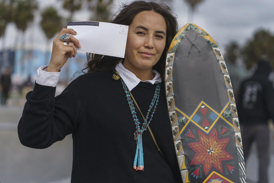 Expert skateboarder Di'Orr Greenwood, an artist born and raised in the Navajo Nation in Arizona and whose work is featured on the new U.S. stamps, holds an art gallery postcard waiting for her own skateboard stamp in the Venice Beach neighborhood in Los Angeles Monday, March 20, 2023. On Friday, March 24, the U.S. Postal Service is debuting the "Art of the Skateboard," four stamps that will be the first to pay tribute to skateboarding. The stamps underscore how prevalent skateboarding has become, especially in Indian Country, where the demand for designated skate spots has only grown in recent years. (AP Photo/Damian Dovarganes)