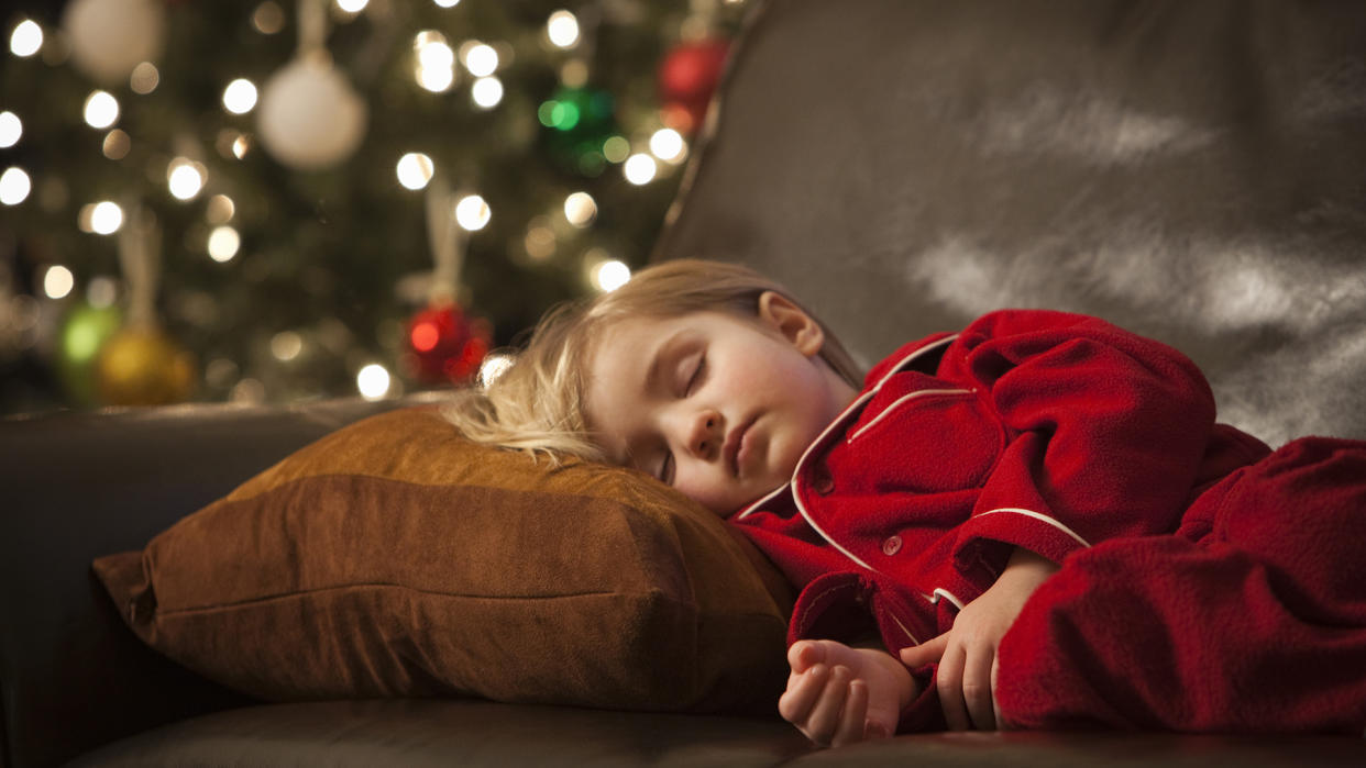  A kid sleeps on the sofa with a Christmas tree in the background. 
