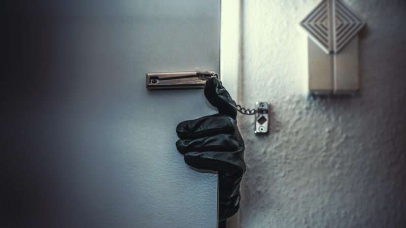 A gloved hand reaches through a cracked door that still has the deadbolt on.