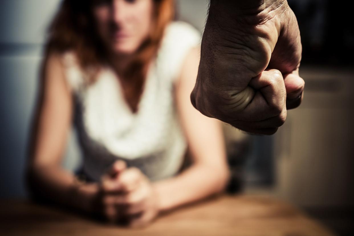 Man's fist in the foreground on the right, he is standing up, with a blurred woman sitting at a table in the background, grey wall, darkness over image