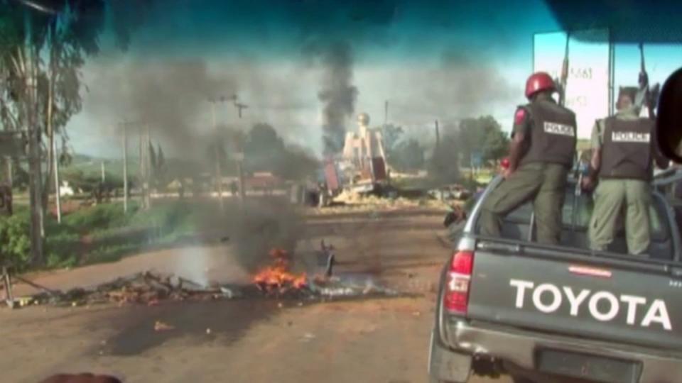 Polizisten passieren in Jos in Nigeria einen Brandherd auf einer Straße. Foto: Nigeria Government/AP