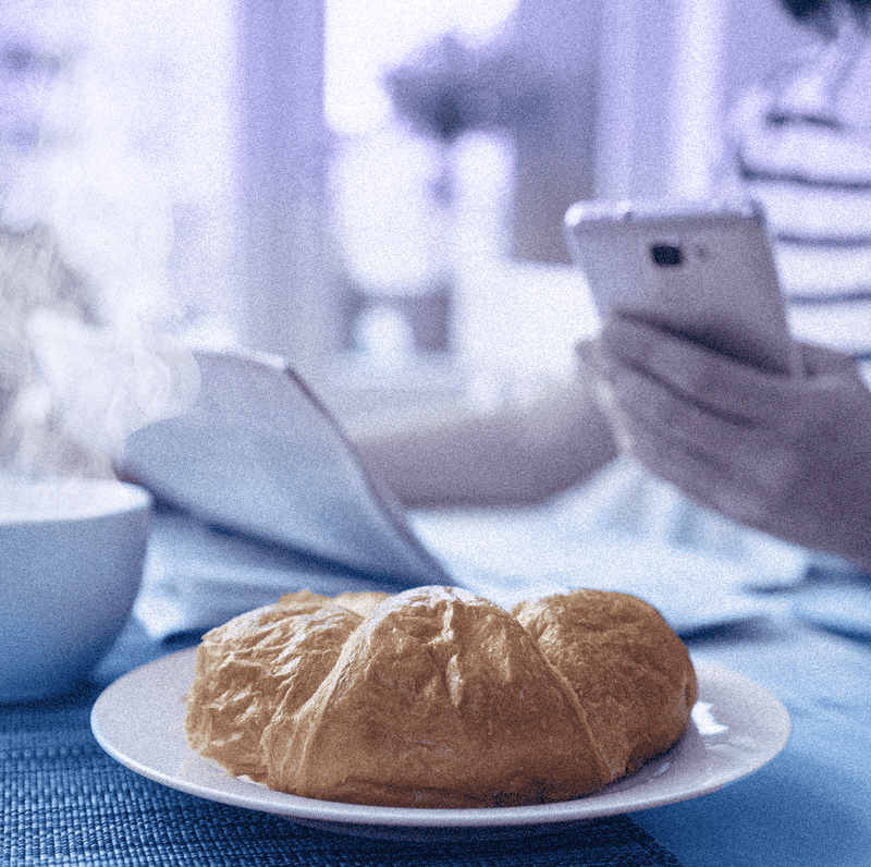 A croissant on a plate with a cup of coffee and a person holding a mobile phone and newspaper