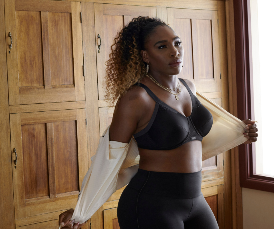 Serena Williams in black leggings and a black sports bra taking off a cream cardigan against a wooden wardrobe.