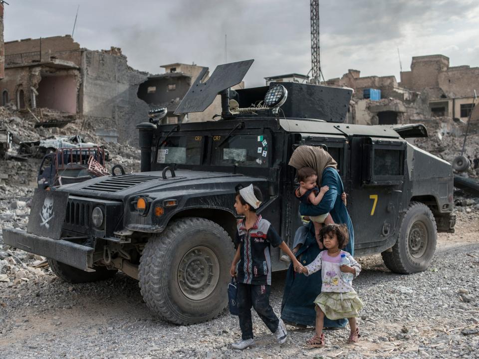 A woman with three children walking in front of a military vehicle in Mosul, Iraq, in front of damaged buildings.