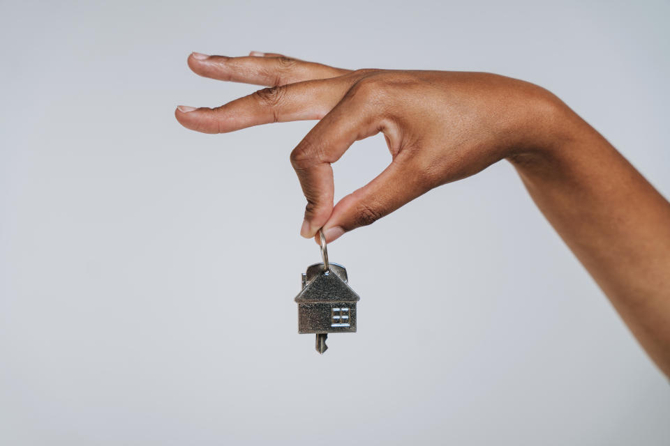 A hand holding a small house-shaped keychain between thumb and index finger against a plain background