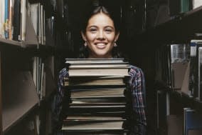 Secondary School Girl Stands Between Bookshelves in a Library Holding a Large Pile of Books