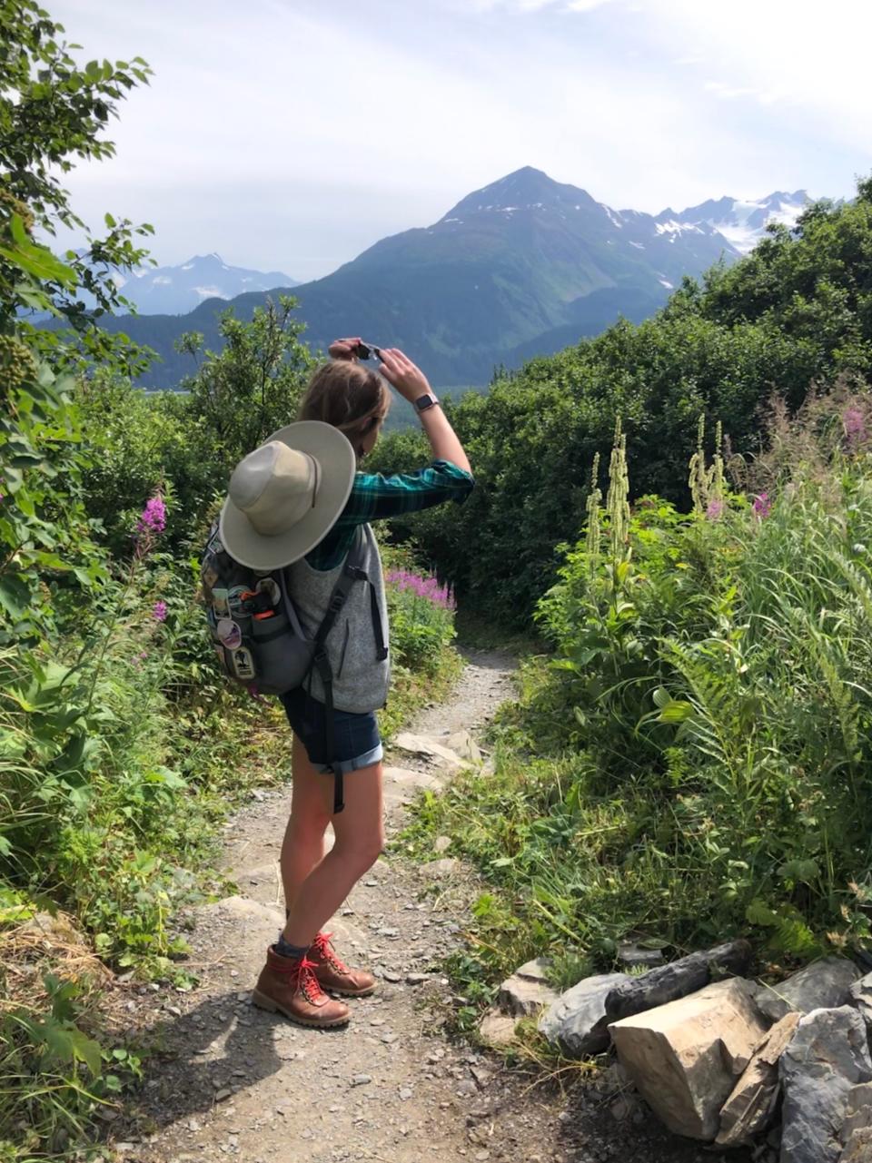 Emily, wearing a hat on her back, hiking boots, shorts, and a vest, looks out towards the mountains on an overgrown trail. 