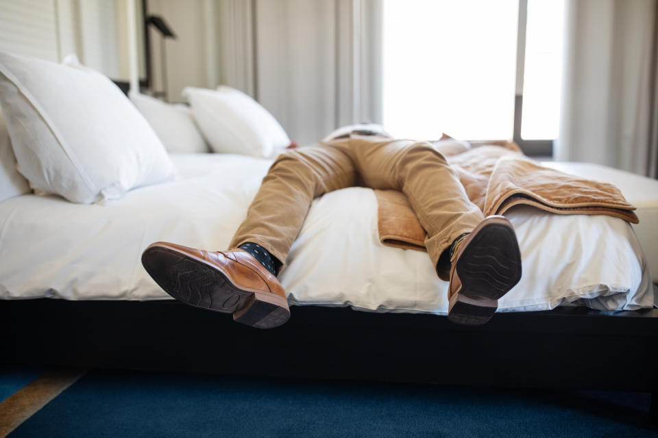 Person lying on a bed with feet crossed, wearing beige pants and brown shoes, indicating exhaustion or break from work