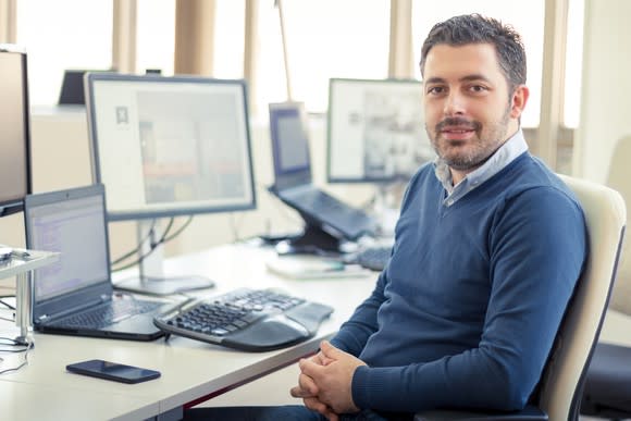 Man sitting at desk smiling with multiple computer screens in the background.