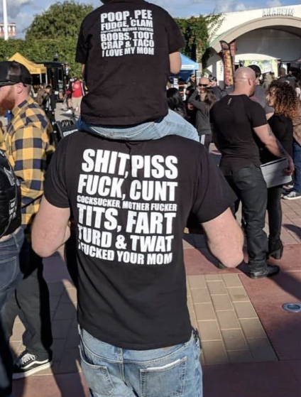 Child on parent's shoulders; both wear profanity-laden shirts at an event. Shirt text reflects bond and humor